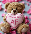 A plush tan teddy bear holding a pink embroidered heart with you are cute af from cheeky chappies bear collection